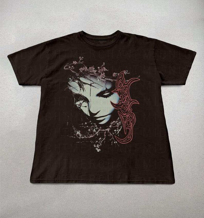 K&ouml;nnte beinhalten: Dunkelbraunes T-Shirt mit einer Grafik eines stilisierten Gesichts, floralen und abstrakten Elementen. Das Gesicht ist in Blau und Wei&szlig; gehalten, mit roten Akzenten. Das Design ist mittig auf der Vorderseite des T-Shirts platziert.
