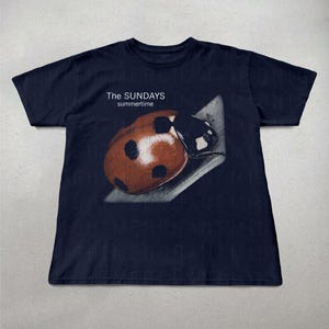 Pode incluir: T-shirt azul marinho com uma imagem de uma joaninha e o texto "The Sundays summertime". A joaninha &eacute; castanha com manchas pretas. Uma camiseta casual para uso di&aacute;rio.