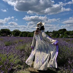Op de afbeelding: Een persoon draagt een lange, bedrukte kimono met paarse bies en een strohoed met strik, staand in een lavendelveld. De kimono heeft een crème- en paars ikat-ontwerp. De lucht is blauw met pluizige witte wolken.
