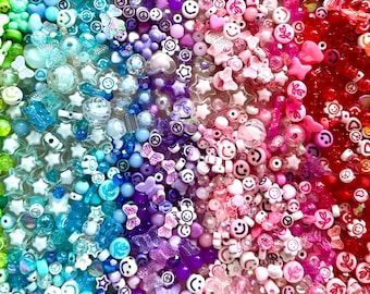 Puede incluir: Una gran colección de cuentas para manualidades variadas dispuestas en un degradado de colores del arcoíris. Las cuentas presentan diversas formas, incluyendo caras sonrientes, estrellas, corazones, mariposas y flores en tonos amarillo, verde, azul, morado, rosa, rojo y naranja.