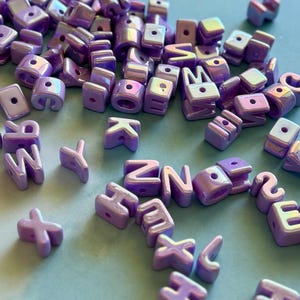 May include: A collection of scattered, iridescent purple alphabet beads, each featuring a single hole for threading. The beads have a smooth, glossy finish with a pearlescent sheen, displaying various letters of the alphabet in a block font style.