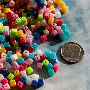 May include: A collection of colorful, square beads in various shades of red, pink, blue, green, yellow, and white. A U.S. dime is included for size reference. The beads are scattered on a light blue surface.