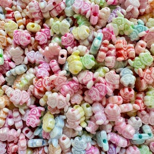 May include: A close-up shot of a pile of colorful beads in various shapes, including butterflies, turtles, and other whimsical designs. The beads are in pastel shades of pink, yellow, green, and blue, with a glossy finish. These beads are ideal for jewelry making and crafting.
