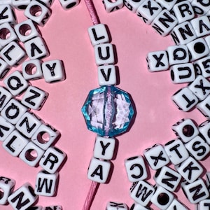 May include: A pink cord with white letter beads spelling "LUV YAY" and a large, faceted blue bead. The beads are surrounded by scattered letter beads on a pink background, creating a playful and colorful design.