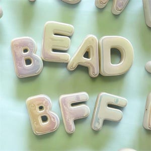 May include: A collection of individual, three-dimensional, iridescent white letter beads arranged to spell the words BEAD and BFF. Each letter has a smooth, rounded, balloon-like shape with a glossy finish that reflects light in soft pastel tones.