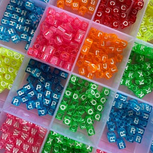 May include: A clear plastic organizer filled with colorful alphabet beads. The beads are in various colors, including blue, orange, red, pink, and green. Each bead has a white letter on it, perfect for crafting personalized jewelry or accessories.