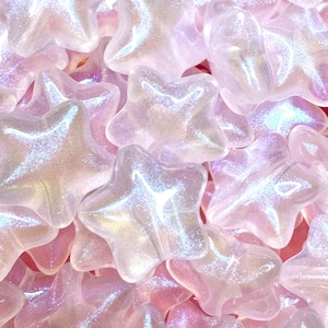 May include: A close-up shot of a pile of iridescent star-shaped beads. The beads are a mix of pink, white, and lavender, with a shimmering, pearlescent finish. The stars are clustered together, creating a textured, decorative effect.