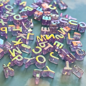 May include: A collection of iridescent purple alphabet beads scattered across a surface. The beads are transparent with a holographic effect, displaying various letters and numbers. Ideal for jewelry making or crafting projects.