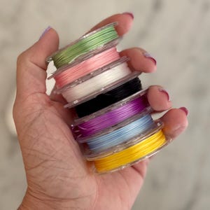 May include: A stack of seven small, clear plastic spools wound with thin, colorful cord. The colors from top to bottom are light green, soft pink, white, black, vibrant purple, light blue, and bright yellow, all held together in a vertical column.