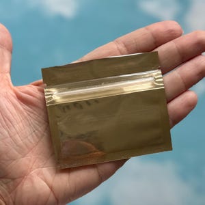 May include: A small, square, metallic gold pouch featuring a clear horizontal window across the center and a resealable zipper closure at the top. The pouch has a reflective, shiny finish and is designed for storage of small items or samples.