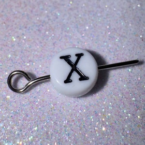 May include: White round bead with the letter X printed in black. The bead is attached to a silver wire with a loop at one end.