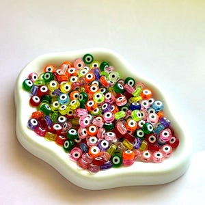 May include: A white, shallow dish filled with a variety of colorful glass beads. The beads are primarily red, orange, green, and blue, each featuring a white and black eye design. The beads are small and translucent, creating a playful and vibrant aesthetic.
