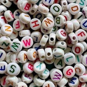 May include: A collection of white, round beads with colorful letters. Each bead has a different letter in a variety of colors, including red, blue, green, and orange. The beads are scattered, showing their holes for stringing.