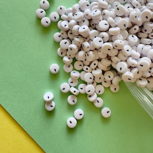 May include: A collection of white, round beads with a black dot on each. The beads are scattered on a green and yellow surface. The beads appear to be made of plastic or a similar material, and are likely used for jewelry making or crafts.