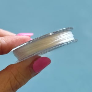 May include: A spool of clear, elastic beading cord. The cord is tightly wound on a transparent plastic spool. The background is a solid light blue.