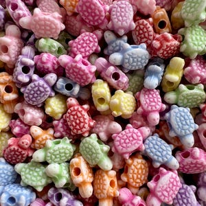 May include: A close-up shot of a collection of colorful turtle-shaped beads. The beads are in various pastel colors, including pink, blue, green, orange, and yellow. Each bead has a hole for stringing.