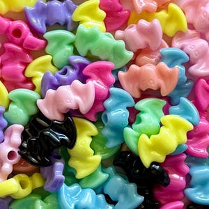 May include: A collection of small, glossy, bat-shaped beads in a variety of pastel and vibrant colors including pink, yellow, purple, green, blue, orange, and black. Each bead features a central hole for threading and a molded bat silhouette.