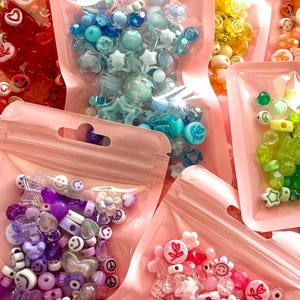 May include: Assortment of colorful beads in clear, resealable pink plastic bags. Beads include various shapes like stars, flowers, smiley faces, and hearts in shades of blue, purple, pink, yellow, and green. Ideal for jewelry making and crafting projects.