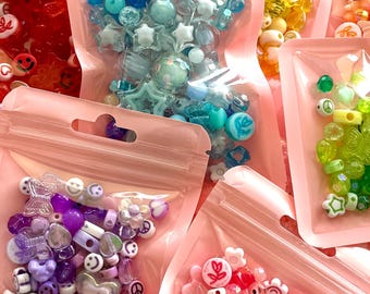 Puede incluir: Variadas cuentas de colores en bolsas de plástico rosa transparente y resellables. Las cuentas incluyen varias formas como estrellas, flores, caras sonrientes y corazones. Los colores incluyen tonos de azul, morado, rosa, amarillo y verde. Ideal para hacer joyas y manualidades.