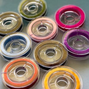 May include: A collection of colorful thread spools in various shades, including pink, green, orange, and purple. The spools are transparent, showcasing the thread wound around the central core. The threads appear to be for crafting or sewing projects.