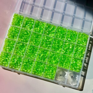 May include: A clear plastic organizer filled with bright green beads. The beads are contained in individual compartments, suggesting organization for crafting or jewelry making. Two spools of clear string are visible in the bottom right corner.
