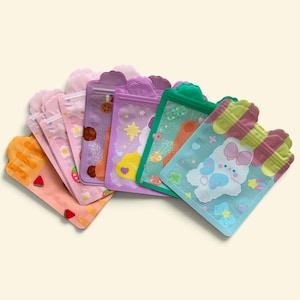 May include: A collection of rectangular resealable pouches with scalloped tops, featuring various pastel colors and holographic finishes. The designs include printed graphics of fruits, stars, clouds, and a white character with a pink bow.
