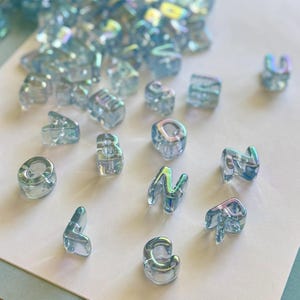 May include: A collection of iridescent blue alphabet beads. The beads are transparent and have a subtle rainbow sheen. The letters are in a variety of fonts and sizes, perfect for jewelry making or craft projects.