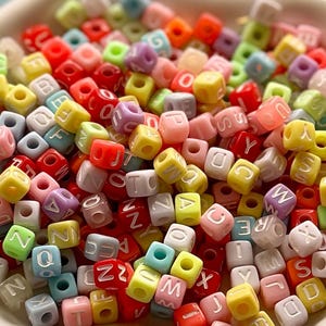 May include: A collection of colorful, square alphabet beads in a white bowl. The beads are in various colors, including red, yellow, pink, green, and blue. Each bead has a letter of the alphabet printed on it in white. Ideal for jewelry making and crafts.