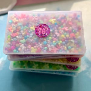May include: A stack of three clear plastic containers filled with colorful beads and embellishments. The top container has a pink sticker that says "BEAD BFF". The beads are various shapes and sizes, including flowers and stars.