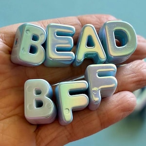May include: Iridescent blue and silver letter beads spelling "BEAD BFF". The beads have a glossy finish and are held in a hand. The letters are in a sans-serif font, perfect for jewelry making or crafting.