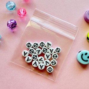 May include: A clear plastic bag filled with white beads featuring black Greek letters. The beads are scattered on a pink surface, alongside colorful, faceted beads and a light blue smiley face bead. The beads are small and round.