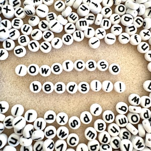 May include: White round beads with black lowercase letters scattered across a beige surface. The words "lowercase beads!" are spelled out with the beads. Ideal for jewelry making and crafting projects.