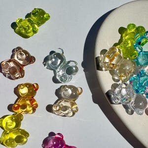 May include: A collection of colorful, translucent gummy bear-shaped charms in various colors, including green, yellow, clear, and pink. Some charms are scattered, while others are arranged on a white, curved surface.