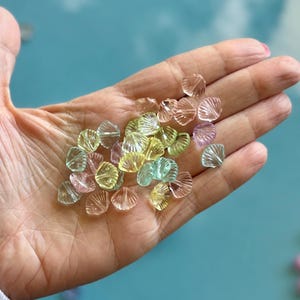 May include: A handful of translucent, seashell-shaped beads in pastel colors: pink, yellow, and green. The beads are scattered across an open palm, with a light blue background. These beads are ideal for jewelry making and craft projects.