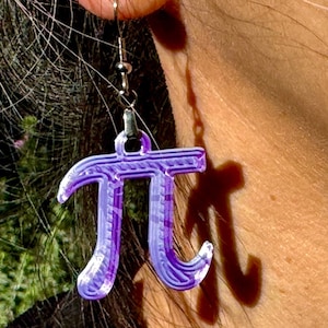 May include: Purple 3D printed pi symbol earrings. The earrings are hanging from silver hooks.