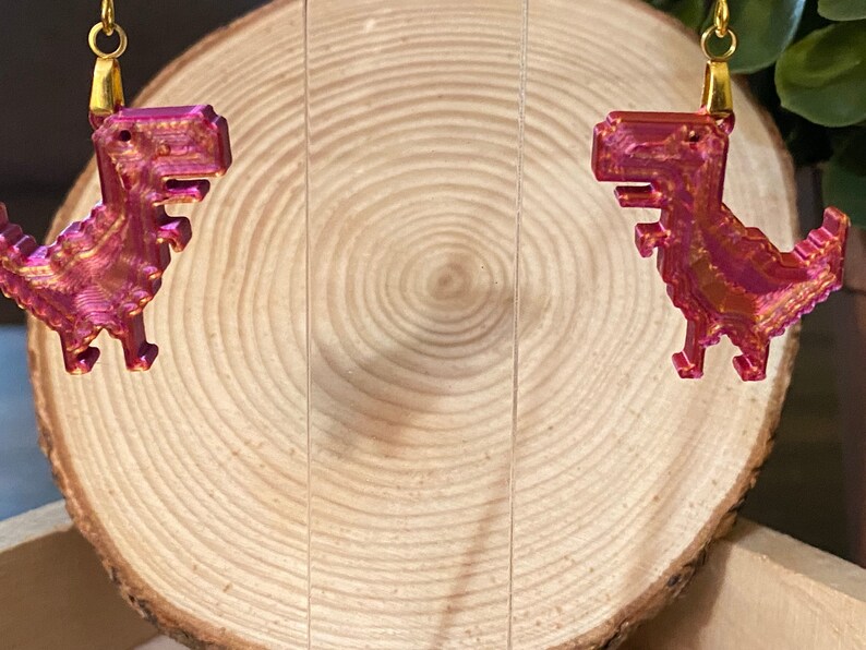 K&ouml;nnte beinhalten: Ein Paar Ohrringe mit einem 3D-gedruckten Dinosaurier-Design in Pink und Gold. Die Dinosaurier sind pixelig und haben einen metallischen Glanz.
