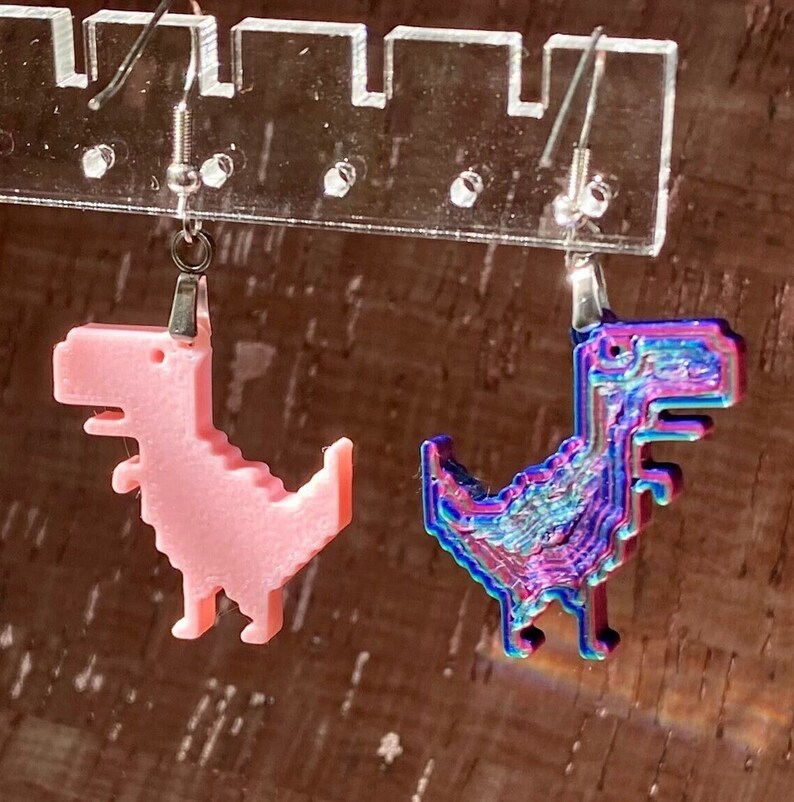 K&ouml;nnte beinhalten: Ein Paar Ohrringe mit einem pinkfarbenen und einem regenbogenfarbenen, pixelartigen Dinosaurier-Design. Die Ohrringe h&auml;ngen an silbernen Haken.
