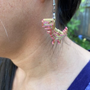 K&ouml;nnte beinhalten: Ein Paar silberne Ohrh&auml;nger mit einem rosa und gr&uuml;nen pixelartigen Dinosaurier-Anh&auml;nger.