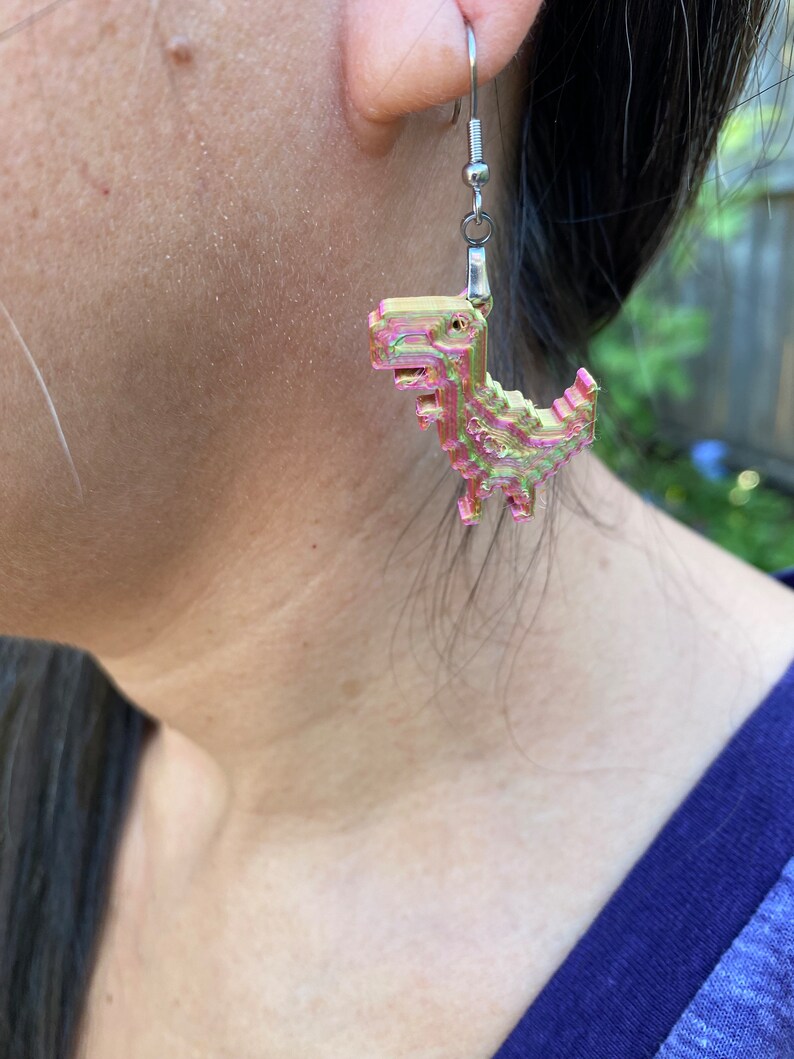 K&ouml;nnte beinhalten: Ein Paar silberne Ohrh&auml;nger mit einem rosa und gr&uuml;nen pixelartigen Dinosaurier-Anh&auml;nger.