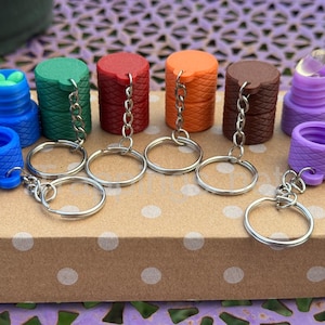 Könnte beinhalten: Eine Sammlung farbenfroher Flaschenverschlüsse mit Schlüsselanhängern. Die Verschlüsse sind in verschiedenen Farben erhältlich, darunter Blau, Grün, Rot, Orange, Braun und Lila. Jeder Verschluss hat ein einzigartiges Design und ist an einem silbernen Schlüsselanhänger befestigt.