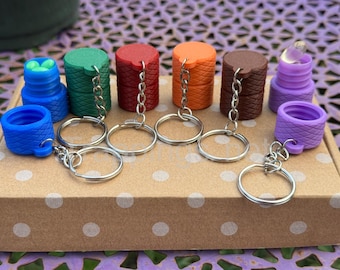 May include: A collection of colorful bottle stoppers with keychains. The stoppers are in various colors, including blue, green, red, orange, brown, and purple. Each stopper has a unique design and is attached to a silver keychain ring.