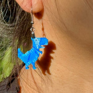K&ouml;nnte beinhalten: Blaue Dinosaurier-Ohrringe mit silbernen Haken. Der Dinosaurier ist ein Pixel-Design.