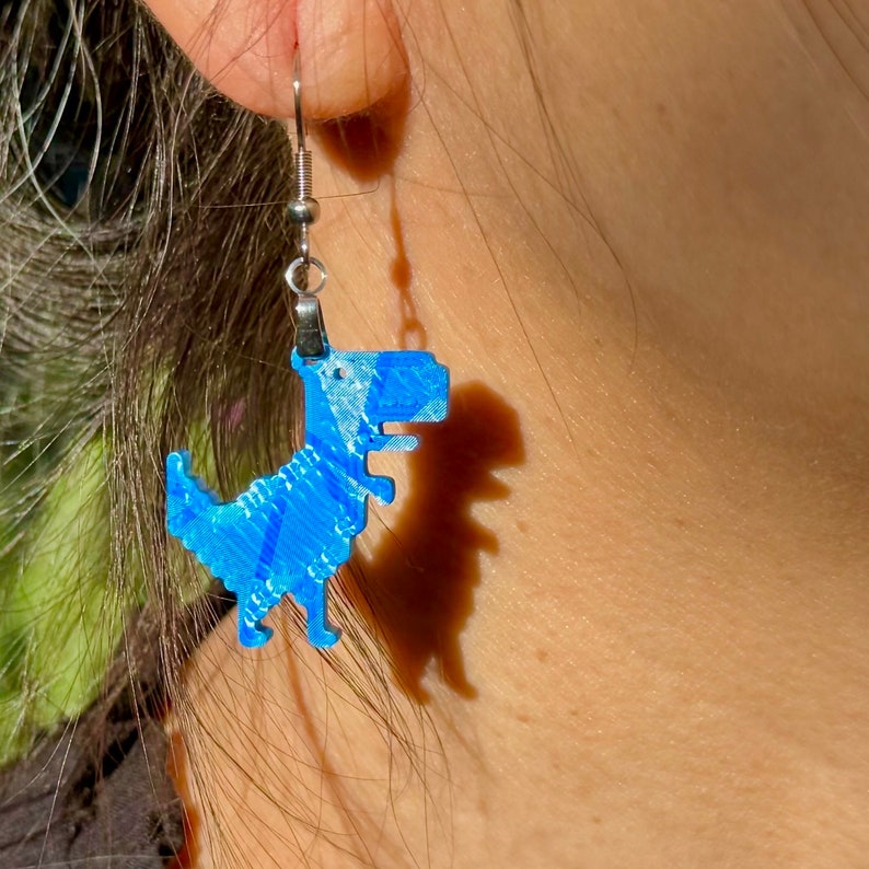 K&ouml;nnte beinhalten: Blaue Dinosaurier-Ohrringe mit silbernen Haken. Der Dinosaurier ist ein Pixel-Design.