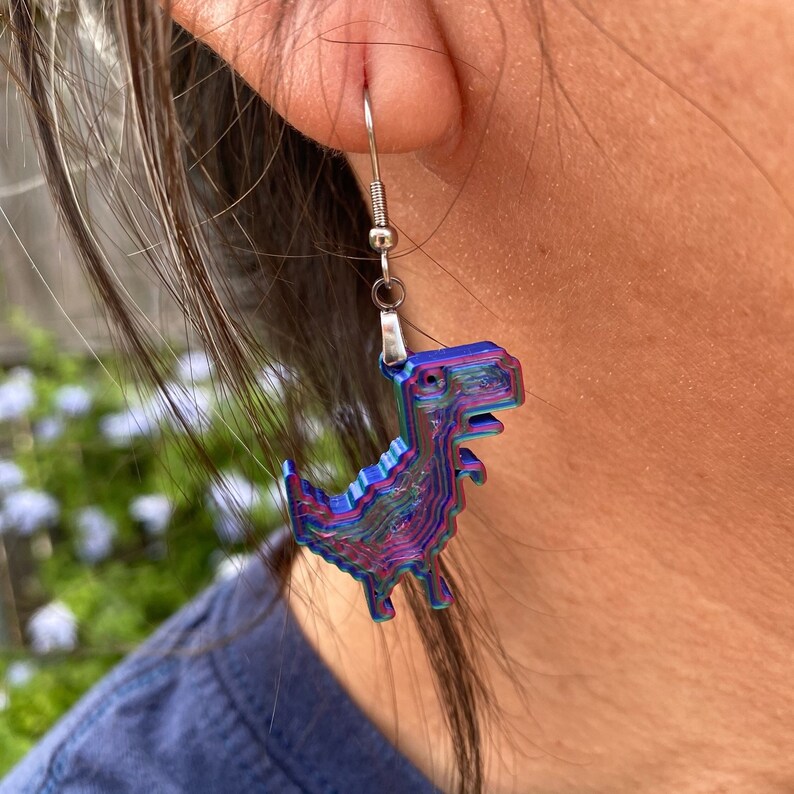 K&ouml;nnte beinhalten: Ein Paar regenbogenfarbene blaue und violette Dinosaurier-Ohrringe. Die Ohrringe sind aus 3D-gedrucktem Material gefertigt und haben ein pixeliges Design.