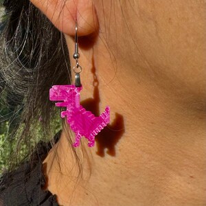 K&ouml;nnte beinhalten: Rosa Dinosaurier-Ohrringe mit silbernen Haken. Der Dinosaurier ist ein Pixel-Design.