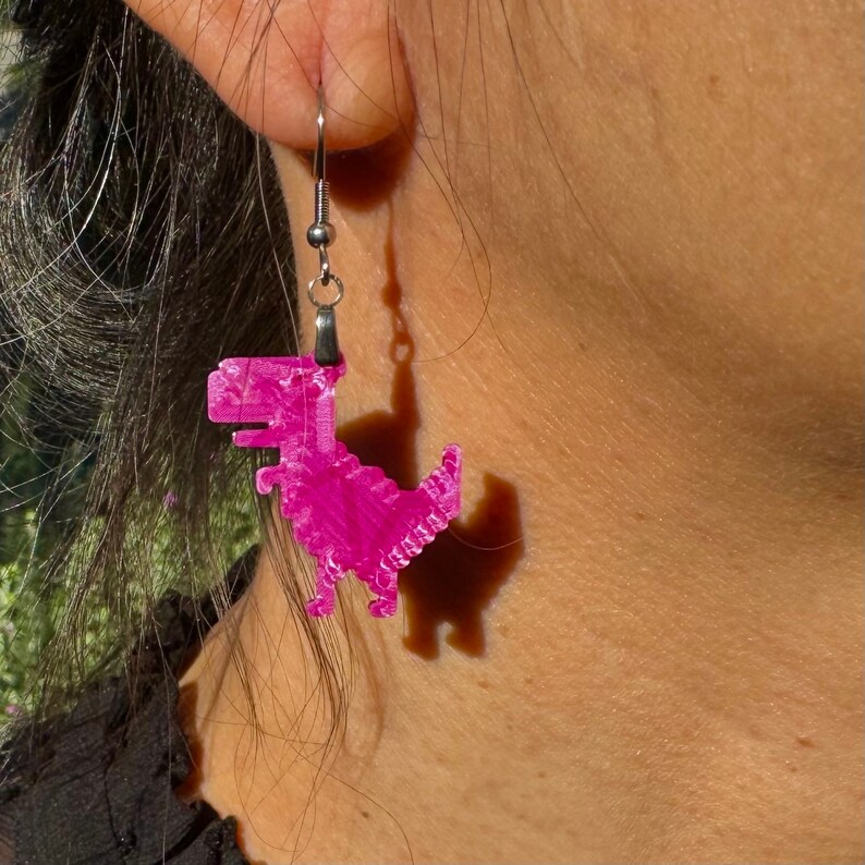 K&ouml;nnte beinhalten: Rosa Dinosaurier-Ohrringe mit silbernen Haken. Der Dinosaurier ist ein Pixel-Design.