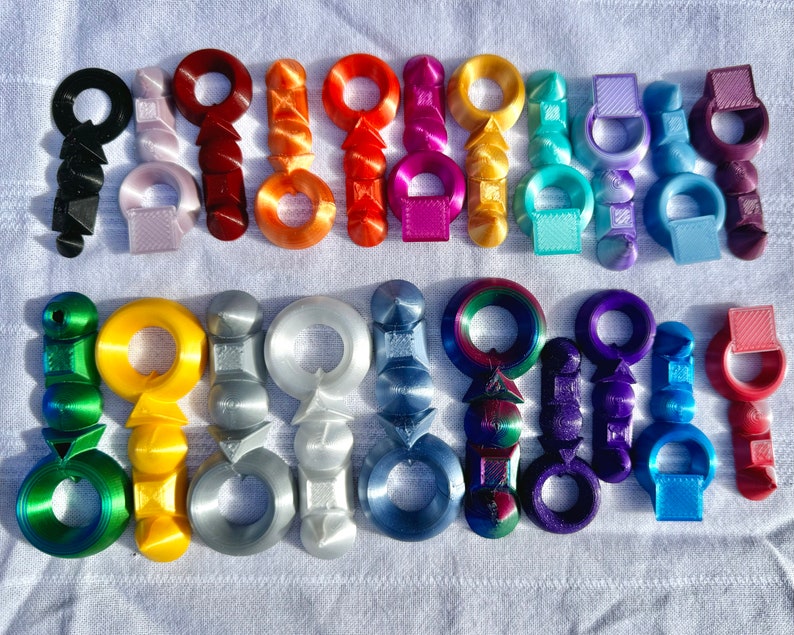 K&ouml;nnte beinhalten: Eine Sammlung von 20 bunten, 3D-gedruckten Schl&uuml;sselanh&auml;ngern. Jeder Schl&uuml;sselanh&auml;nger hat ein einzigartiges Design mit einem Ring oben zum Anbringen an Schl&uuml;sseln oder Taschen. Die Schl&uuml;sselanh&auml;nger sind aus Kunststoff und in verschiedenen Farben erh&auml;ltlich, darunter Rot, Blau, Gr&uuml;n, Gelb und Lila.