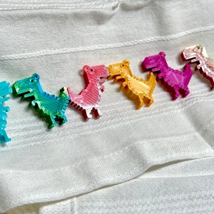 K&ouml;nnte beinhalten: Eine Reihe von sieben bunten, pixelartigen Dinosaurier-Figuren, die in einer Linie auf einem wei&szlig;en Stoffhintergrund angeordnet sind. Die Dinosaurier sind in den Farben des Regenbogens, beginnend mit Blau, Gr&uuml;n, Pink, Gelb, Lila und endend mit einem glitzernden wei&szlig;en Dinosaurier.