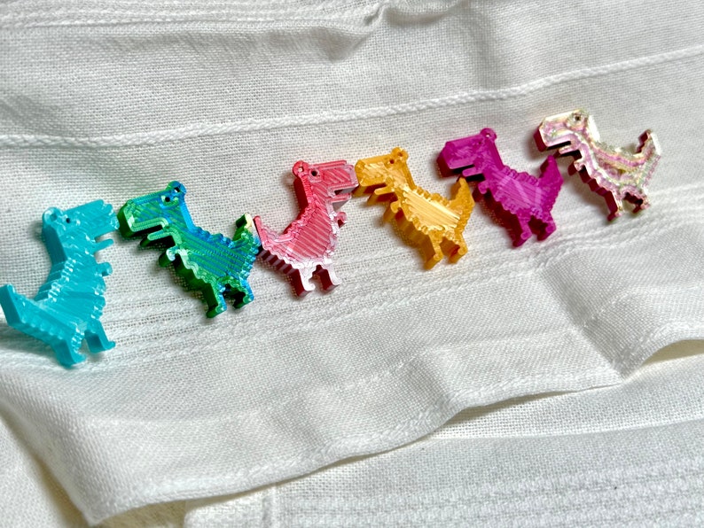 K&ouml;nnte beinhalten: Eine Reihe von sieben bunten, pixelartigen Dinosaurier-Figuren, die in einer Linie auf einem wei&szlig;en Stoffhintergrund angeordnet sind. Die Dinosaurier sind in den Farben des Regenbogens, beginnend mit Blau, Gr&uuml;n, Pink, Gelb, Lila und endend mit einem glitzernden wei&szlig;en Dinosaurier.
