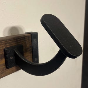 May include: Black metal wall mounted hook with a curved design. The hook is attached to a wooden shelf.